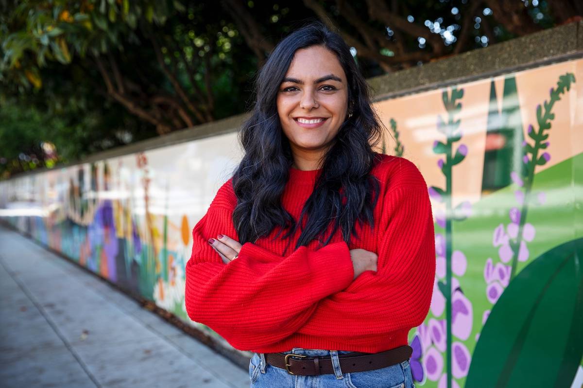 An Indian woman with long dark hair wears a red sweater and stands cross-armed in front of a long mural