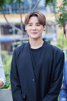 This contain Kim Junsu  standing next to each other in front of trees and bushes, one wearing a black jacket