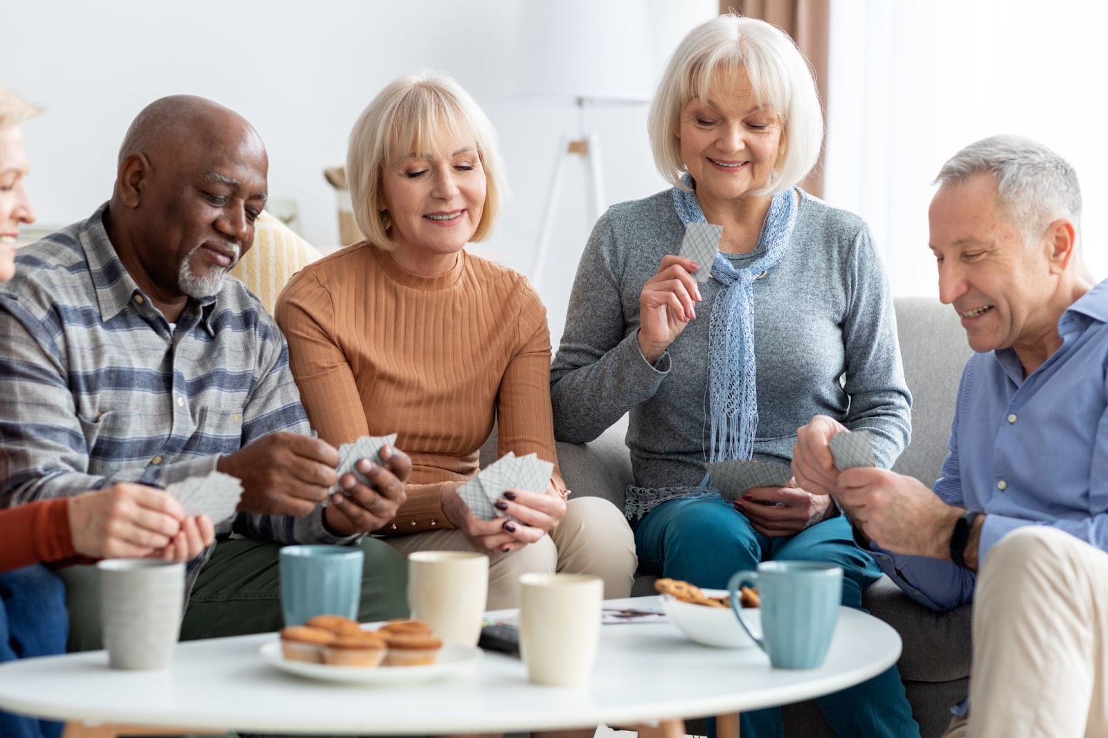 A group of happy seniors sitting around a table eating a snack and playing a game of cards.