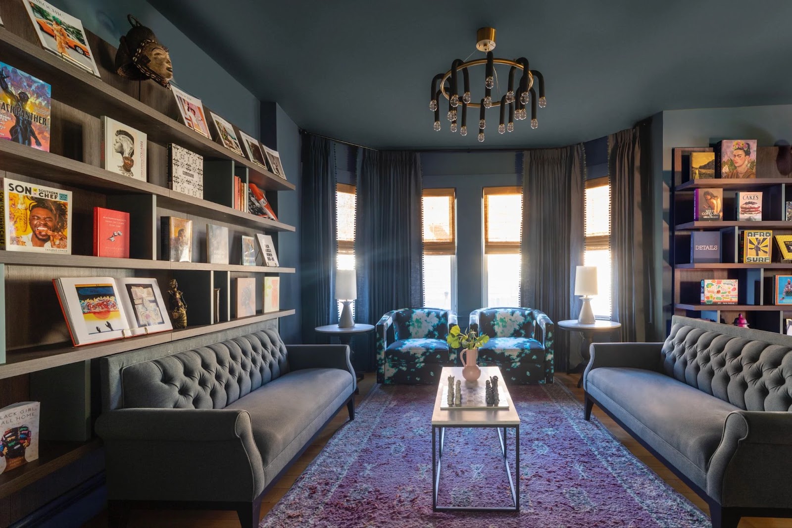 Eclectic dark-toned library lounge with tufted gray sofas, built-in shelves, deep blue walls and ceiling, and a mix of art books and decor pieces.