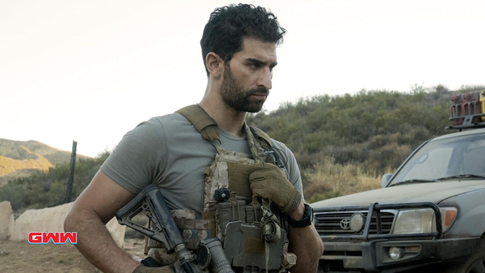 Omar holding a rifle stands near a dusty SUV.