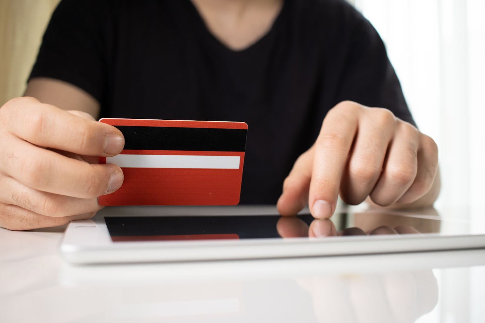 Person holding a red credit card while using a tablet on a white surface