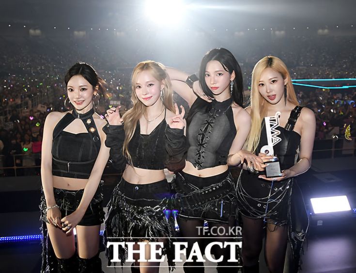 This contain an image of  four girls in black outfits posing for the camera at a concert with an audience behind them