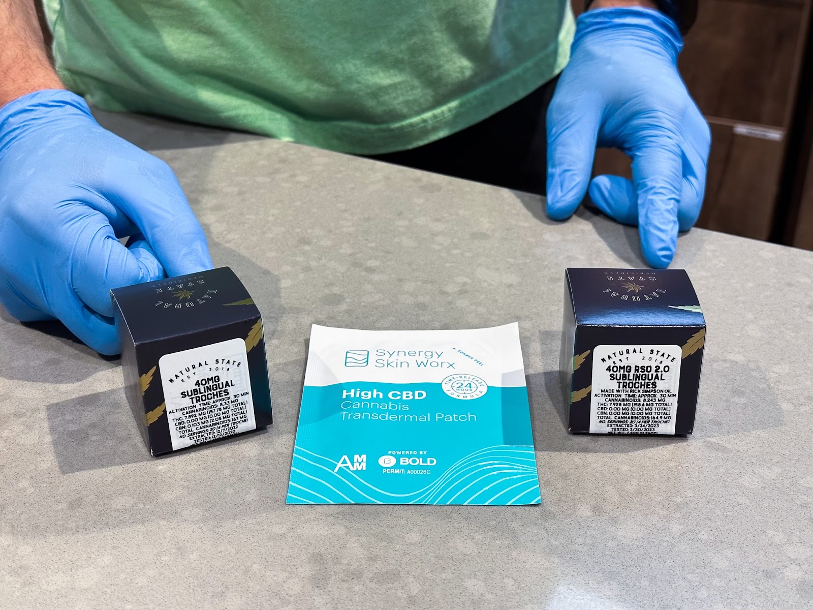 Infused-cannabis sublingual troches by Natural State Medicinals and transdermal patches by Synergy Skin Worx are displayed with a budtender from The Source cannabis dispensary in Rogers, Arkansas in the background.