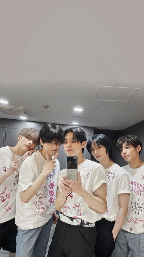 This containTXT members  standing next to each other in front of a mirror looking at a cell phone