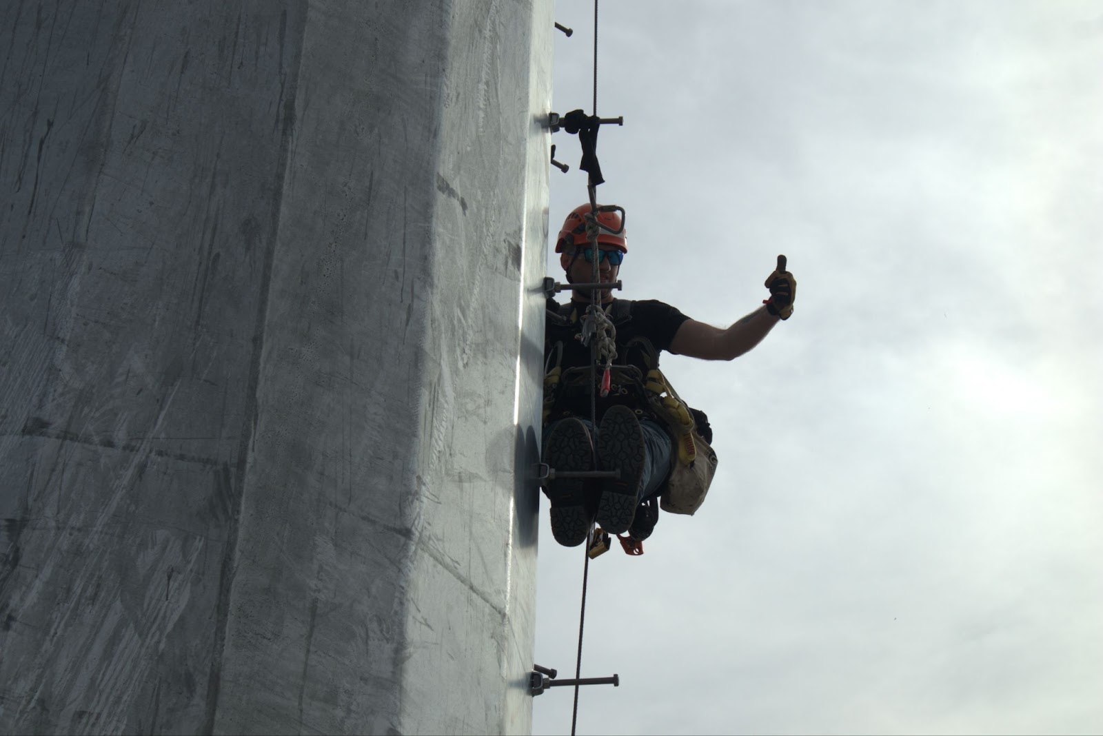 A telecommunications tower technician wearing safety gear, including a helmet, sunglasses, and harness, is suspended on the side of a tower. The technician is holding onto a safety rope with one hand and giving a thumbs-up with the other, displaying confidence and professionalism against a gray, cloudy sky.