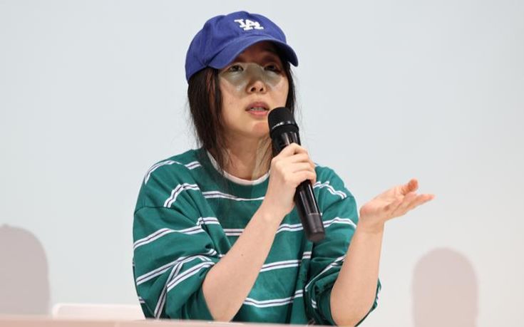 This contain an image of Min Hee Jin holding a microphone in front of her face and wearing a cap on top of her head