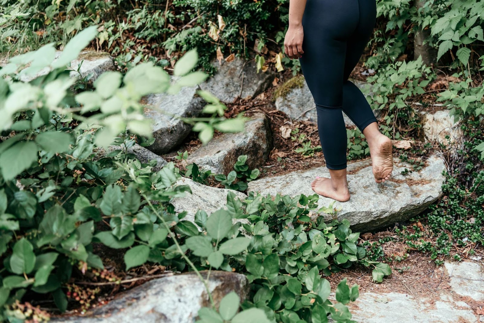 A female figure wearing black leggings and walking barefoot over stones in a woodland setting. 