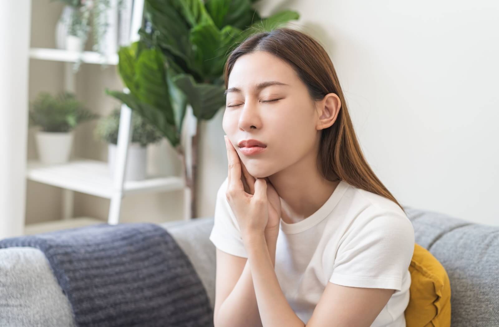 A person sitting on a couch with their eyes closed, holding their sore jaw due to eating acidic food after dental surgery.