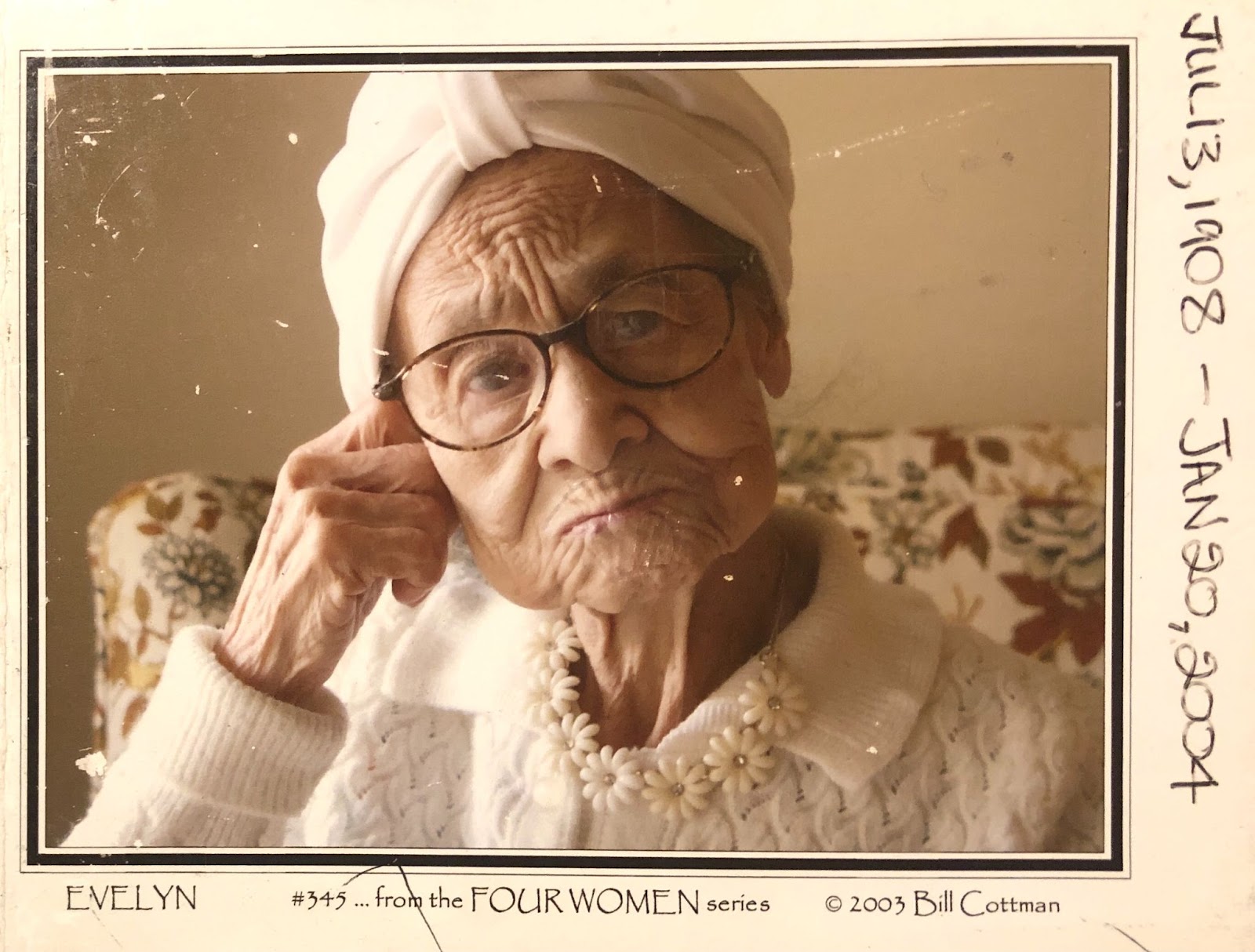 Image: A woman in a white head wrap, sweater, and flower necklace looks center frame. She sits in a floral chair. Evelyn Cottman, William Cottman's mother. Photo courtesy of writer.