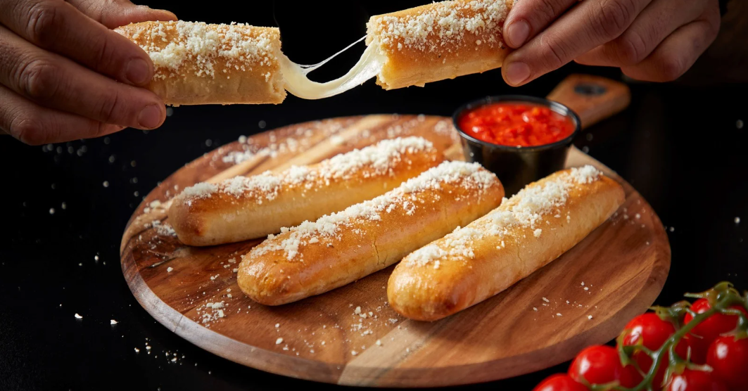(Cheesy garlic breadsticks topped with parmesan and served with marinara dipping sauce, displayed on a wooden board with a stretchy cheese pull in focus.)