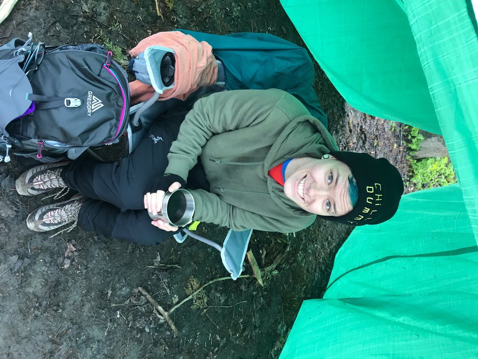 Emory sitting in a camping chair under a tarp holding onto a cup of coffee