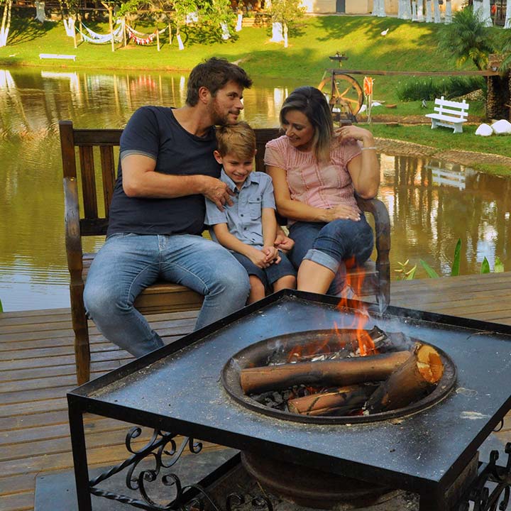Família sorridente aproveitando momento ao ar livre junto à lagoa, sentado em banco de madeira e perto de fogueira em ambiente natural e tranquilo.