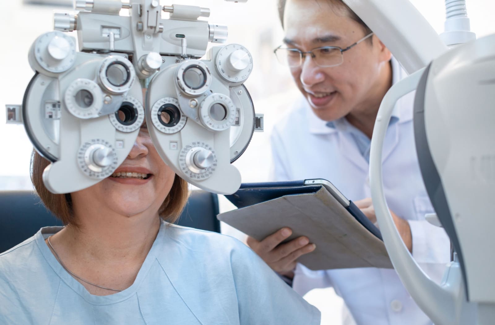 A patient looks through a phoropter during their eye exam while their optometrist evaluates their vision off to the side.