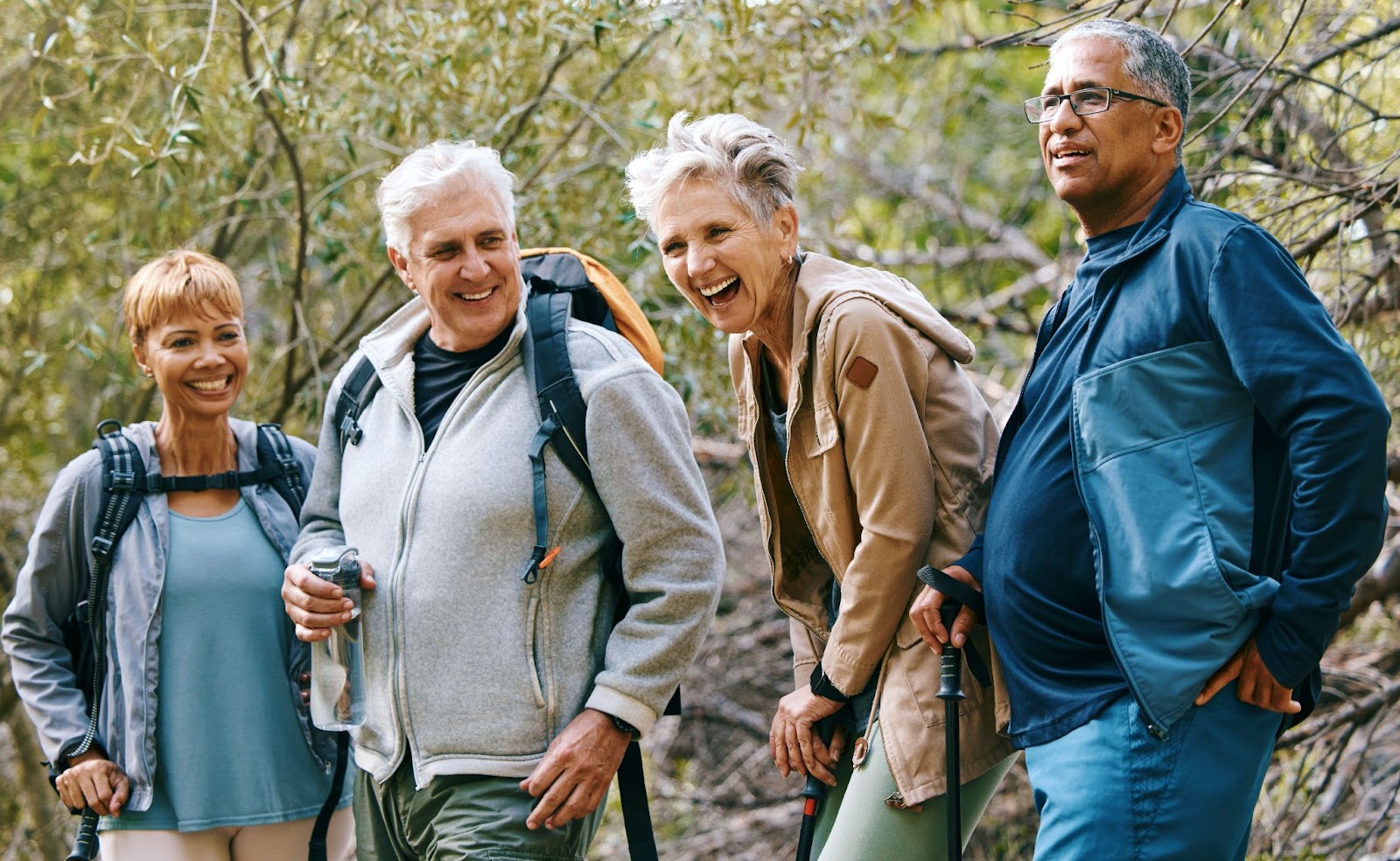 A group of older adults with hiking gear smile and laugh as they go for a hike outdoors.