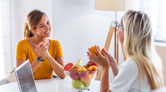 Two women talking around a table with a bowl of fruit, during a nutritional consultation.