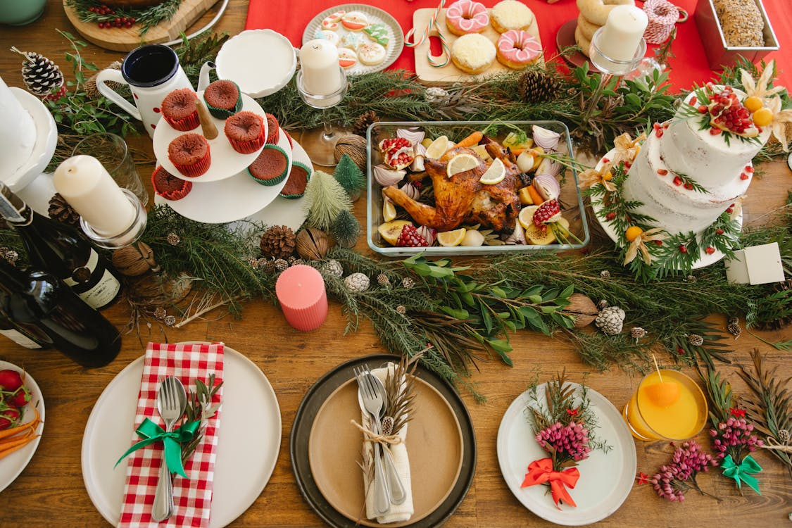 Festive Christmas dinner table decorated with greenery, candles, cupcakes, cookies, a roasted turkey, and a white holiday cake topped with berries and pine.