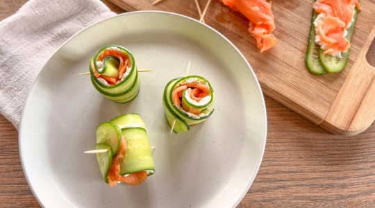 Close-up of cucumber slices rolled with smoked salmon and cream cheese, secured with toothpicks, presented on a white plate.