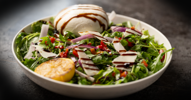 A fresh arugula salad with shaved parmesan, roasted red peppers, red onions, and a ball of burrata, drizzled with balsamic glaze and grilled lemon.