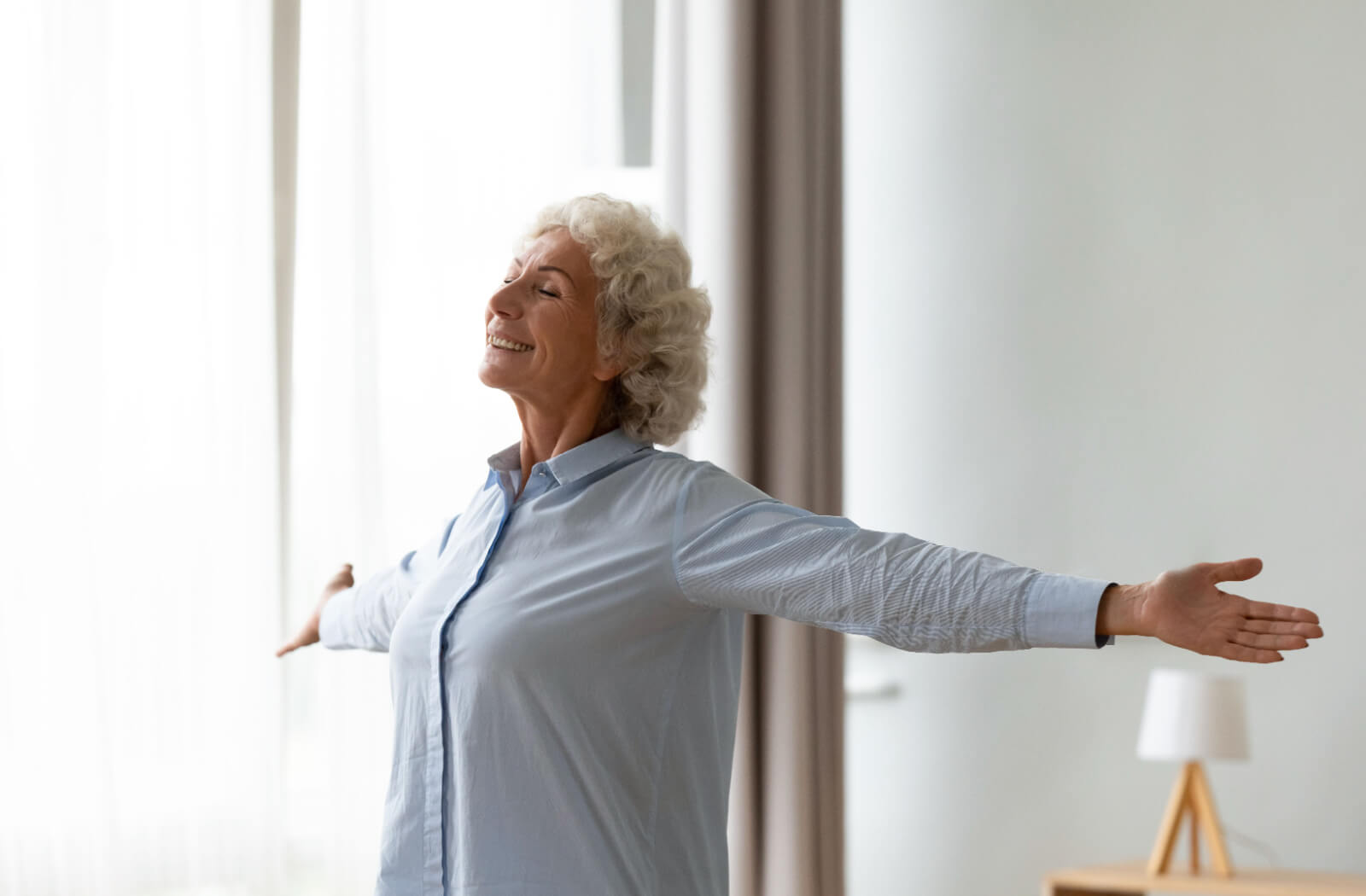 An older adult with their arms outstretched in front of a bright window, smiling in their new senior living villa.