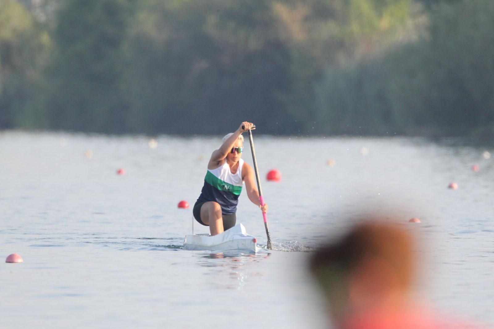 Tarzan paddles a very narrow white canoe in a calm body of water, in a kneeling position on one knee, using a single-bladed paddle to propel the canoe forward. She wears her South Niagara Canoe Club singlet with horizontal green and white stripes and black shorts, and Pit Viper sunglasses. Several buoys are visible on the water's surface, marking the course. In the background is blurred greenery, indicating a shoreline.