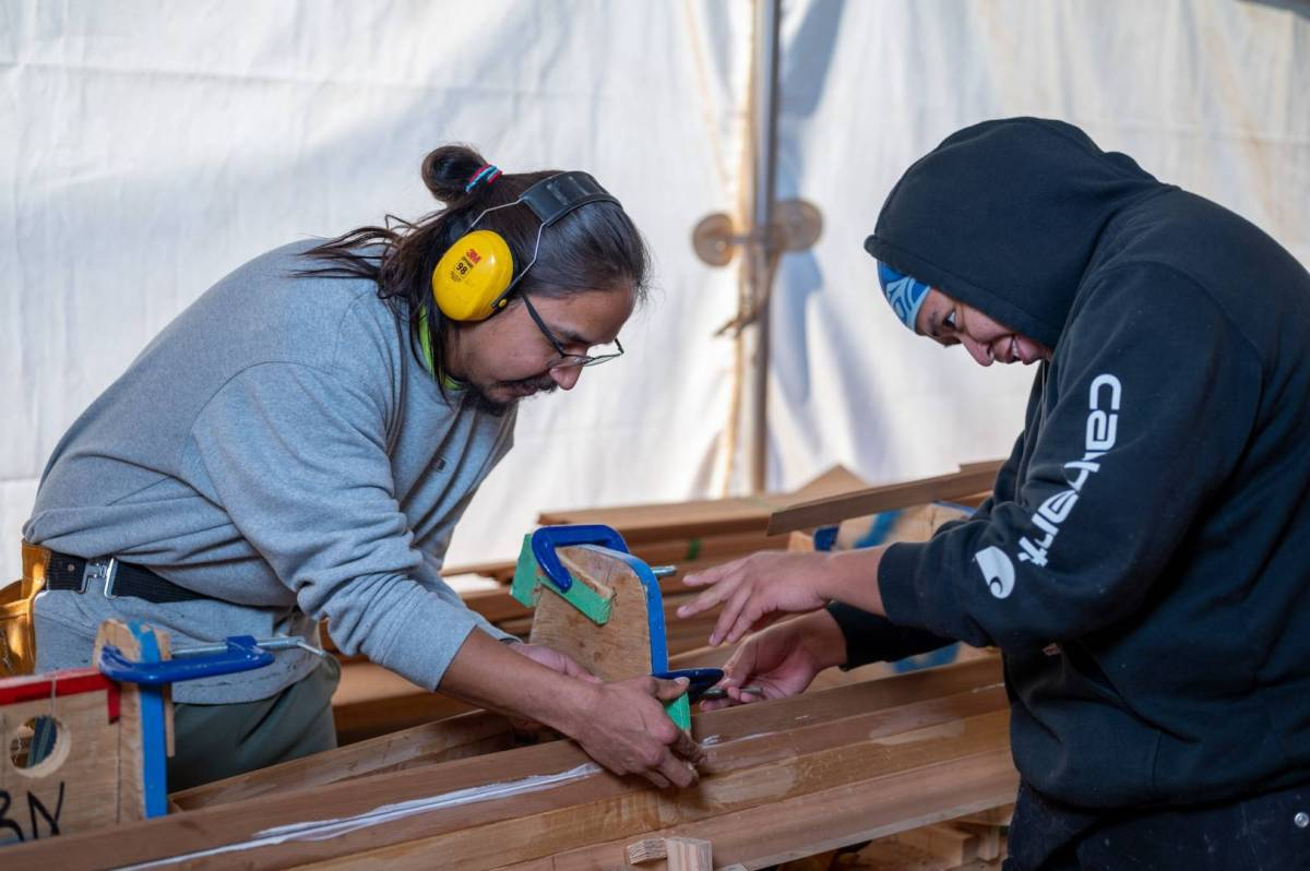 An Indigenous carver demonstrates the skill to an Indigenous youth.