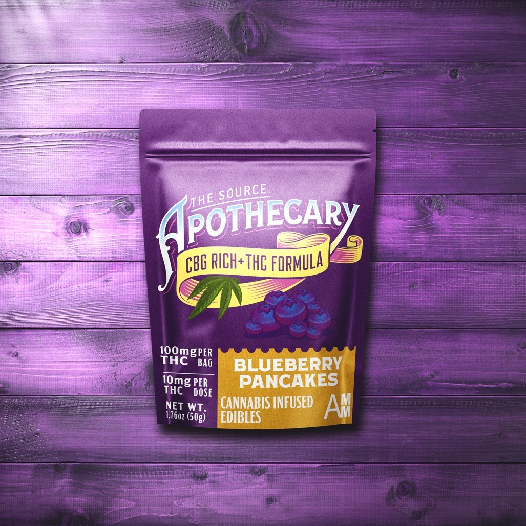 The Source Apothecary Cannabinoid Rich formula Blueberry Pancakes cannabis edibles are enriched with the alternative cannabinoid CBG plus THC. The indigo blue kraft paper packaging is shown from above and the background is multicolored wood.