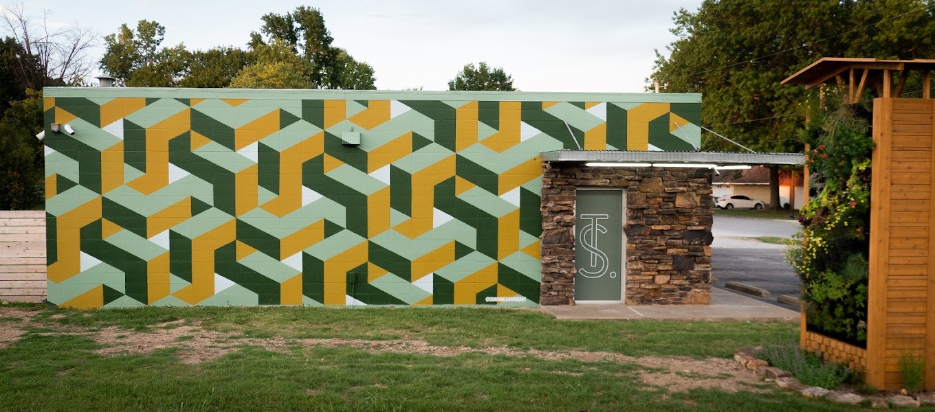 Side view of The Source's Bentonville location from Walton Rd. The building features a striking geometric mural in shades of green and yellow. A stone-faced entrance with The Source's logo on the door is visible, along with part of the 'Living Wall' plant display on the right.