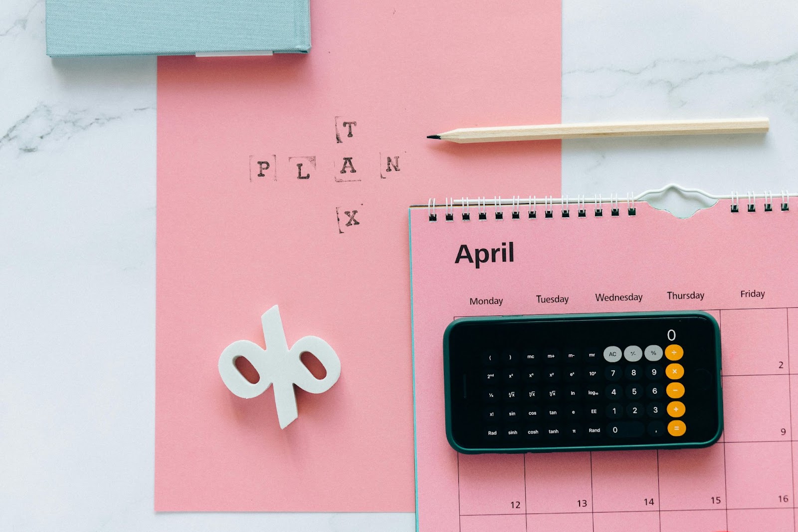 Una calculadora sobre un calendario con un fondo de papel rosa. Se ven también un lápiz, un símbolo de porcentaje y una caja azul