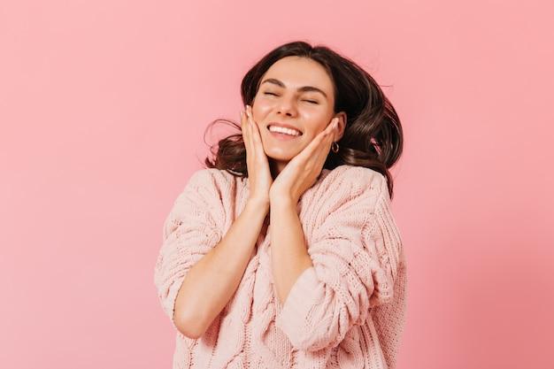 Positive brunette in great mood is laughing and touching her cheeks. Girl in knitted dress posing on pink background.