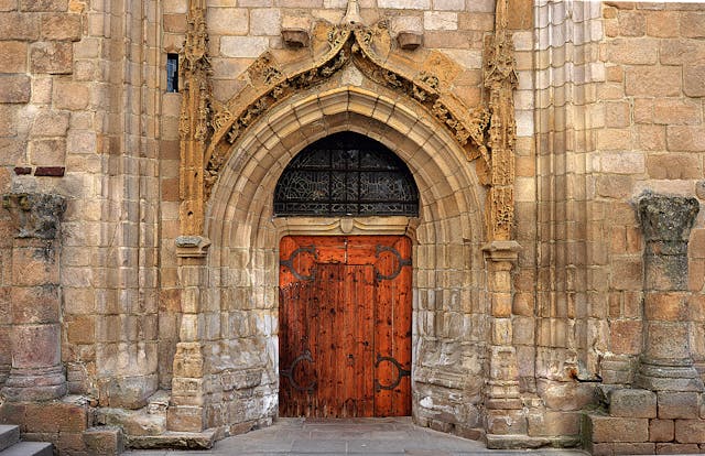 An ornate, arched doorway made of carved stone and dark wood graces a weathered stone building. Reflecting principles of architectural conservation, the intricate medieval design features decorative patterns around the arch and a metal grille above the door.