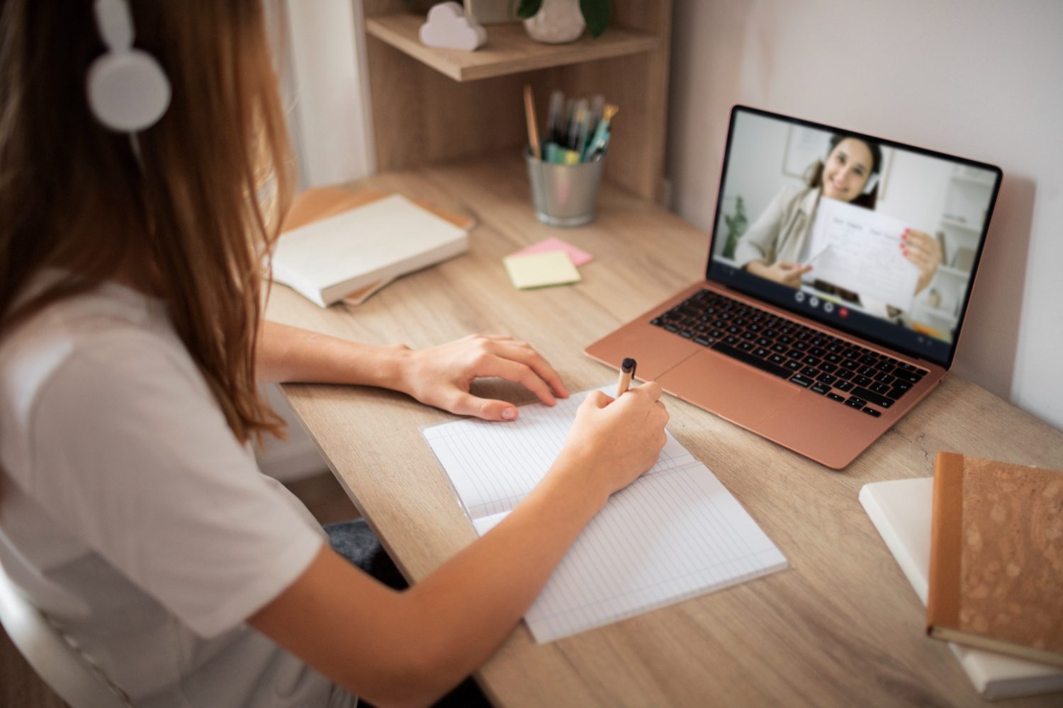 Une jeune fille en train de suivre un cours en ligne à son bureau à domicile. Elle prend des notes dans un cahier tout en écoutant une enseignante visible sur l'écran de son ordinateur portable. 