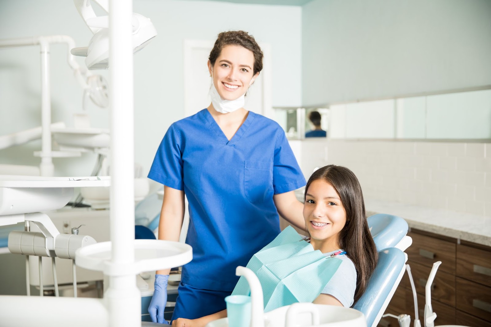 Smiling female dentist in blue scrubs talking to a young female patient seated in a dental chair in a bright, modern clinic—representing professional dental care and patient trust.