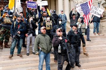 A small group of people stands on the steps of Michigan’s state capitol, holding flags and signs, several armed with guns.