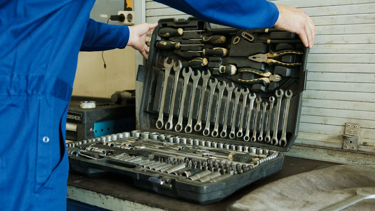 A person in a blue jumpsuit opening a comprehensive toolkit on a workbench, containing a variety of tools such as wrenches, screwdrivers, pliers, and sockets, organized neatly in a black case