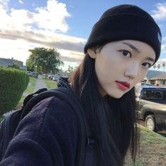 This contains an image of Jung Chae-yul with long black hair wearing a beanie and looking at the camera while standing in front of a car