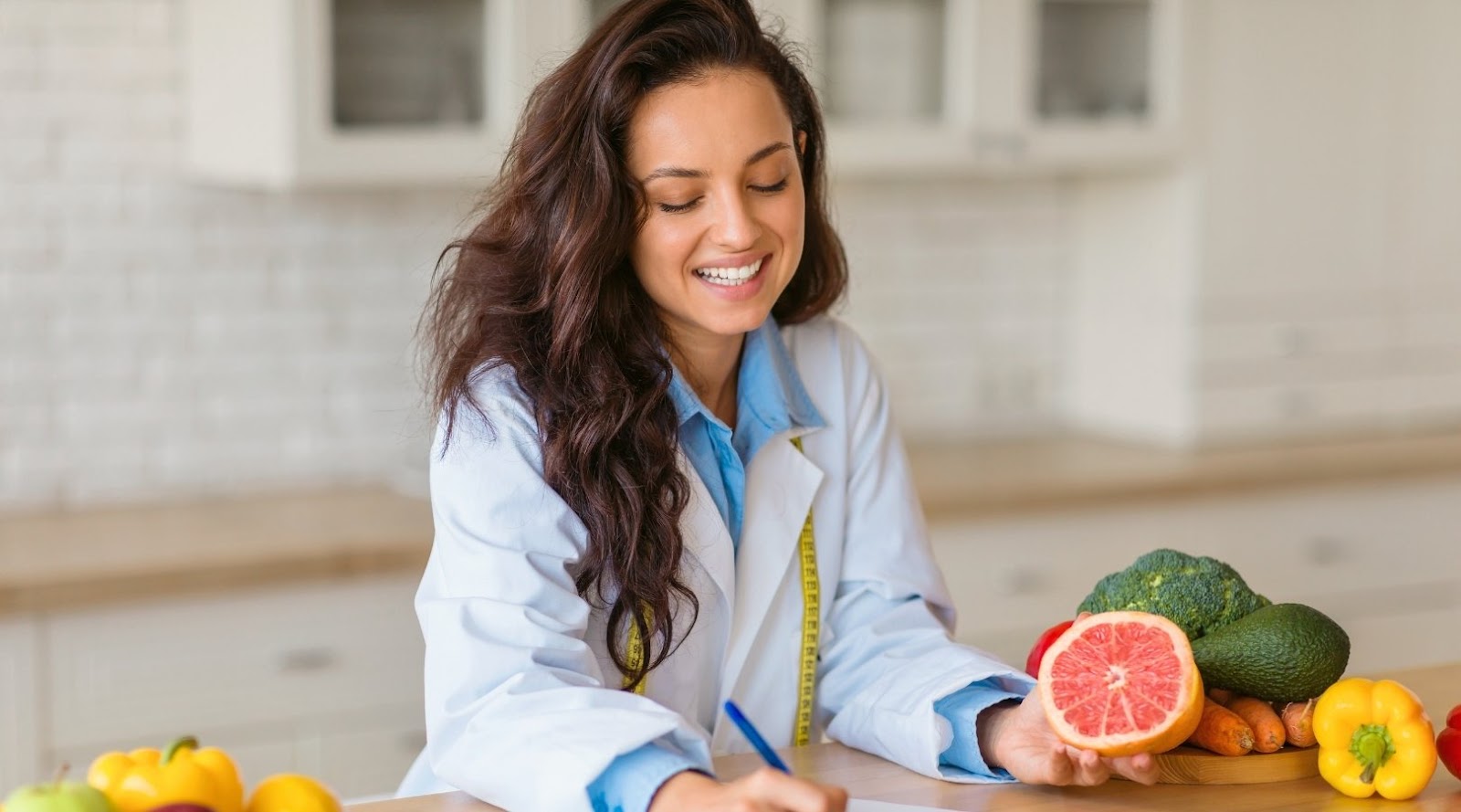 Diététiste-nutritionniste souriante en blouse de laboratoire tenant une moitié de pamplemousse, entourée de légumes frais et prenant des notes.