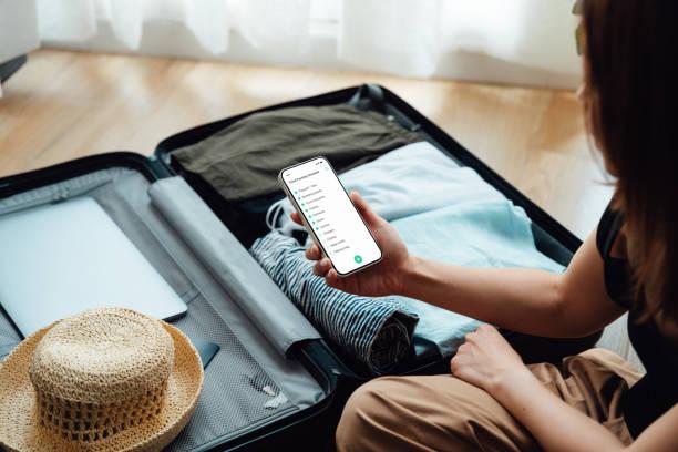 over the shoulder view of young woman looking at checklist on smartphone while packing luggage - checklist stock pictures, royalty-free photos & images