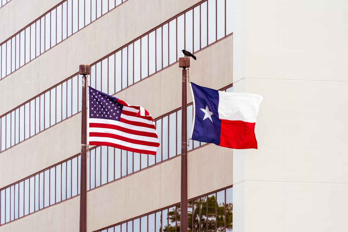 American flag and Texas state flag flying side by side on flagpoles in front of a modern office building, with a black bird perched atop one of the poles.