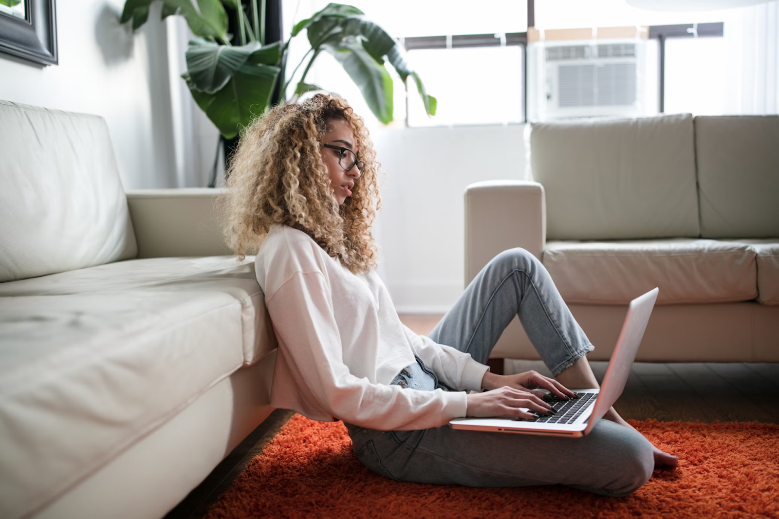 A woman using a laptop while sitting on the floor and leaning against a sofa.