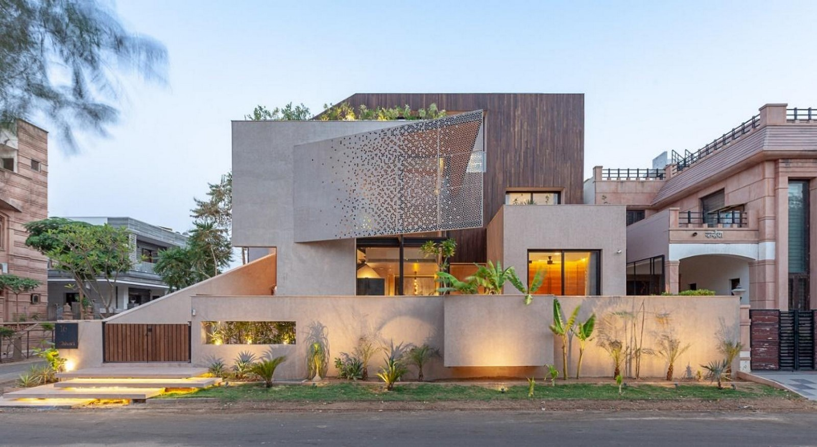 A modern multi-story house designed with vastu principles. It has a geometric design and perforated metal facade, crafted by skilled architects. It features large glass windows, a rooftop garden enhancing energy balance, and a landscaped front yard with plants.