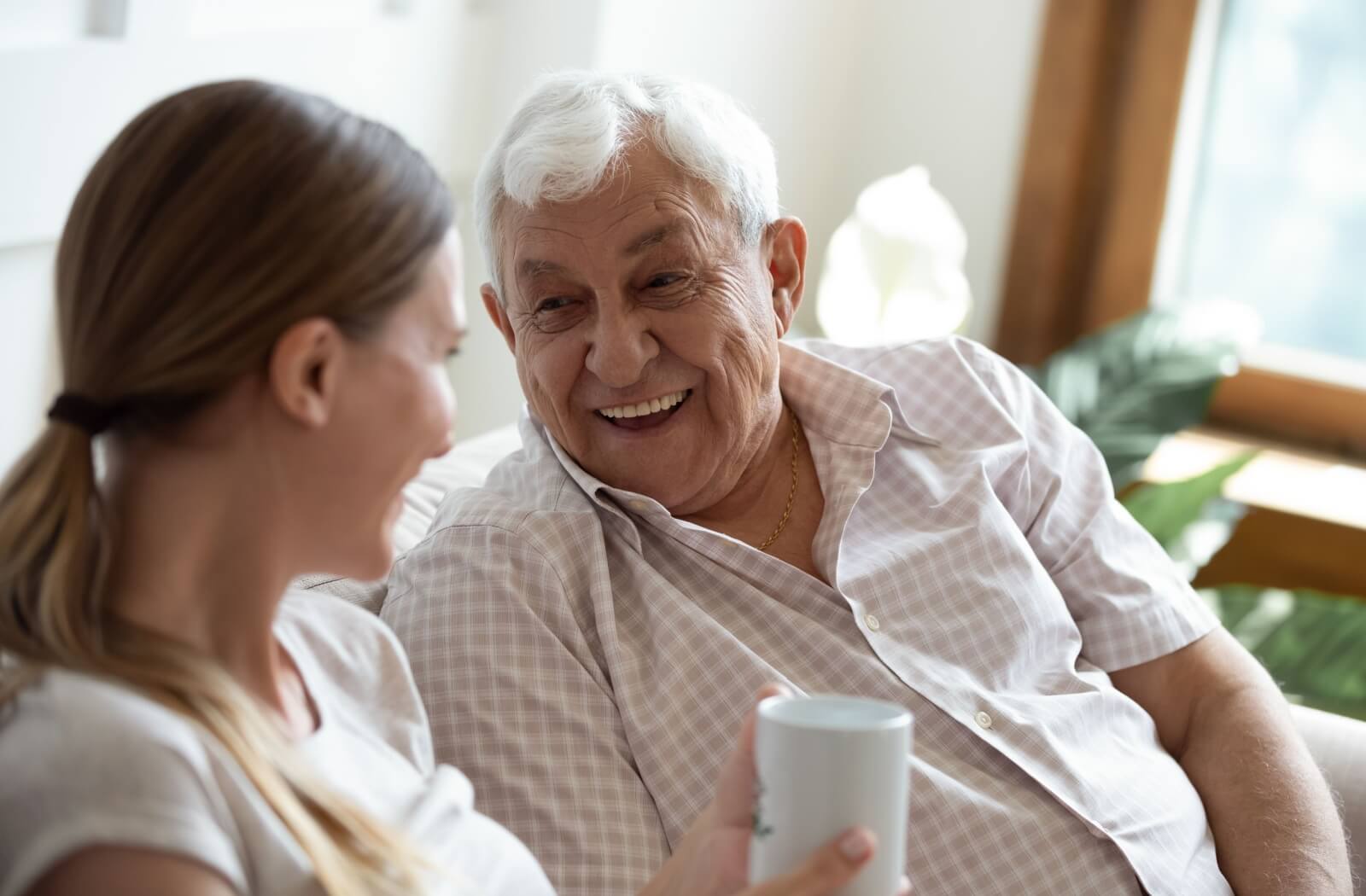 Adult daughter visiting older father and sitting on the couch laughing with him while drinking a cup of coffee.