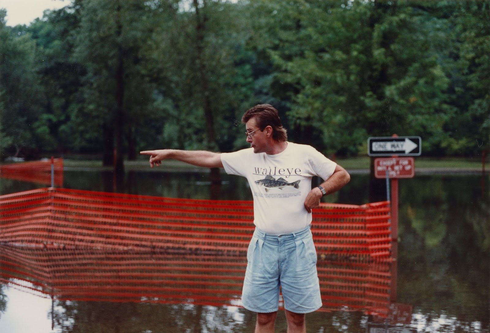 A man stands in front of an orange plastic fence and street flooding while pointing to the left. 