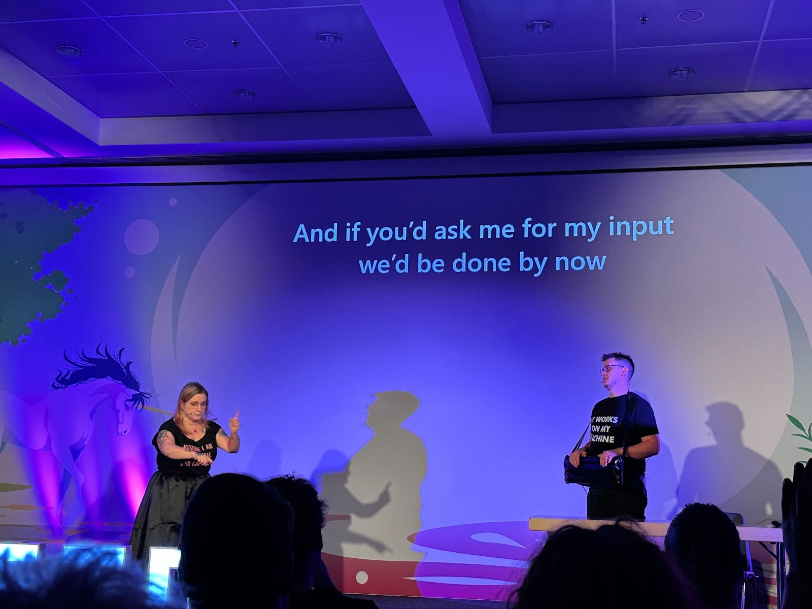 Picture of a stage with a woman and a man standing on it. The test "And if you'd ask me for my input we'd be done by now" is behind them on a screen