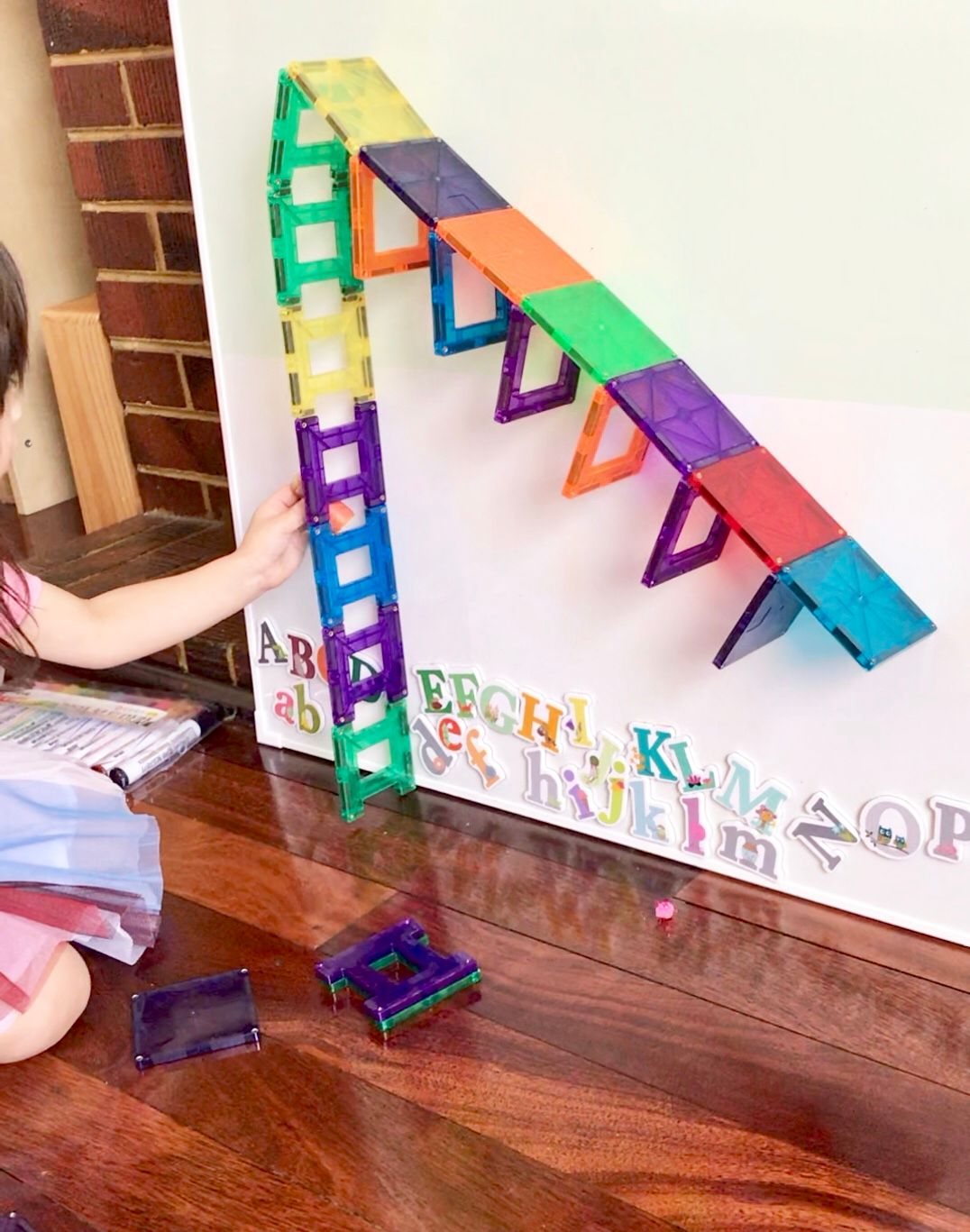 A toddler is lining up colorful magnetic tiles on a magnetic board surface, arranging them to resemble a ladder and a slide. The child's focused play encourages creativity and fine motor skills while exploring STEM concepts such as magnetism, structure, balance, and design.
