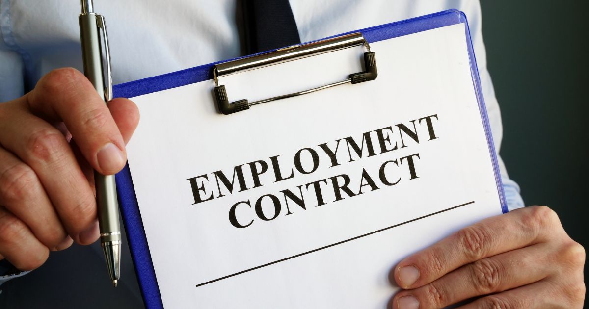 A person holding a clipboard with the document titled "EMPLOYMENT CONTRACT" prominently displayed. The individual is also holding a pen, suggesting readiness to sign or review the contract. The image conveys themes of employment, legal agreements, and formal hiring processes, particularly in the context of Emiratisation or workforce integration.