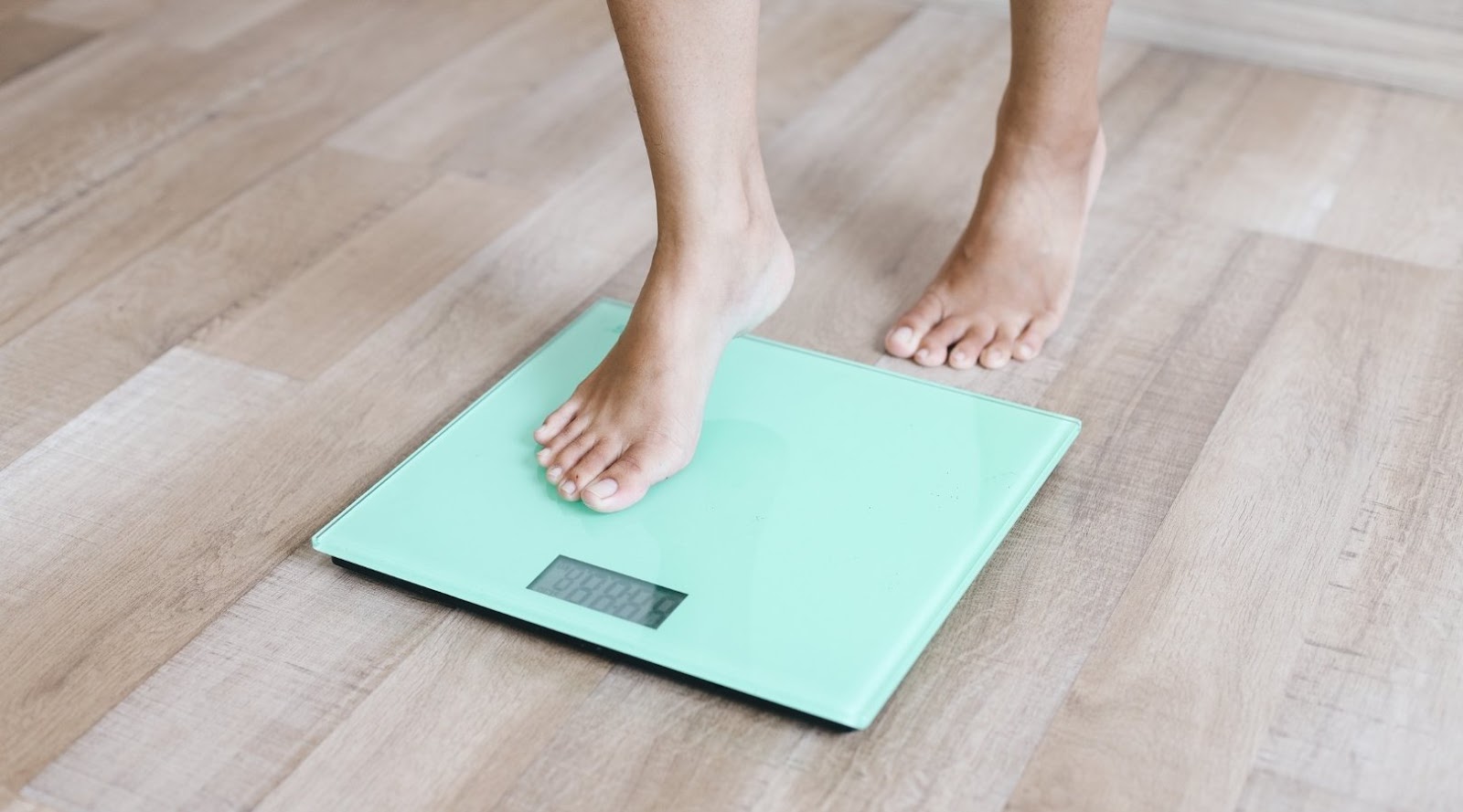 Person stepping onto a green digital scale on a wooden floor, with part of one foot placed on the scale.