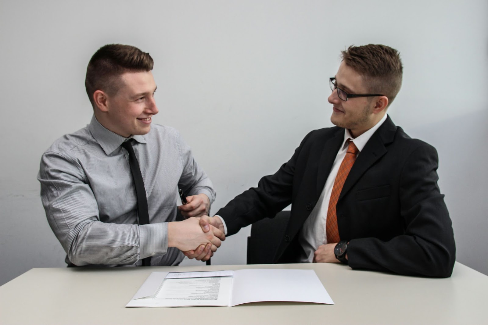 Two businessmen shaking hands across a table, with a document and pen in front of them.