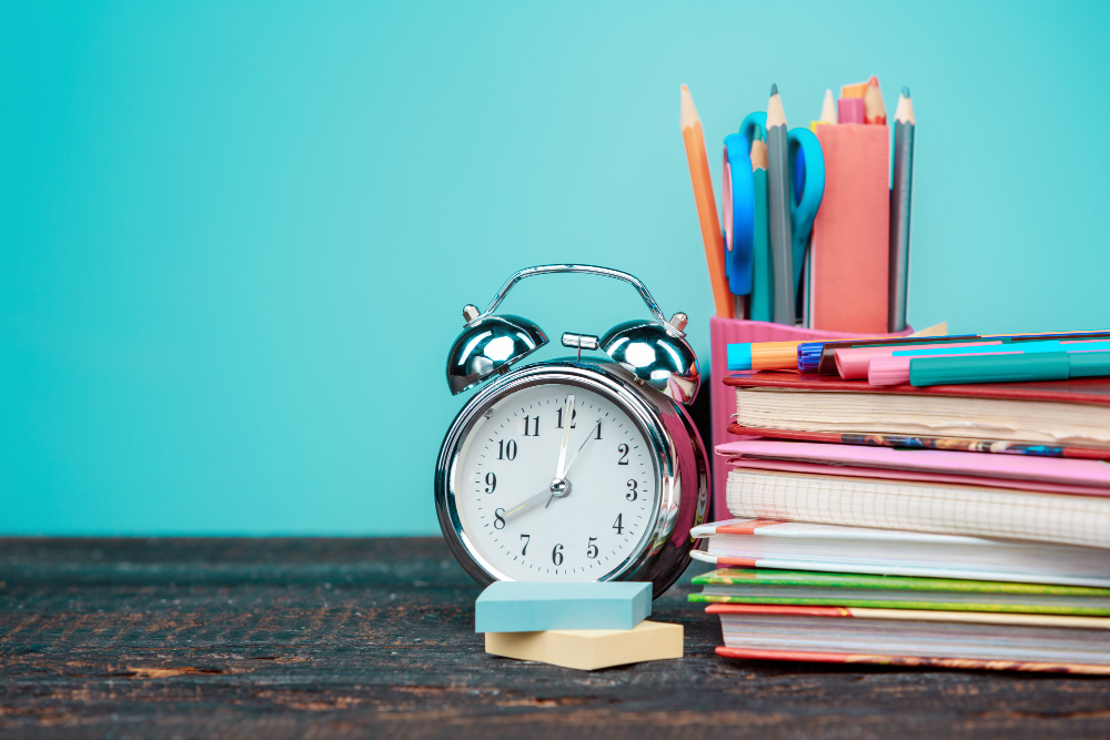 A silver analog alarm clock rests on a wooden surface beside stacks of colorful notebooks and a container filled with pencils, markers, and scissors, serving as the perfect setup for organizing study tips.