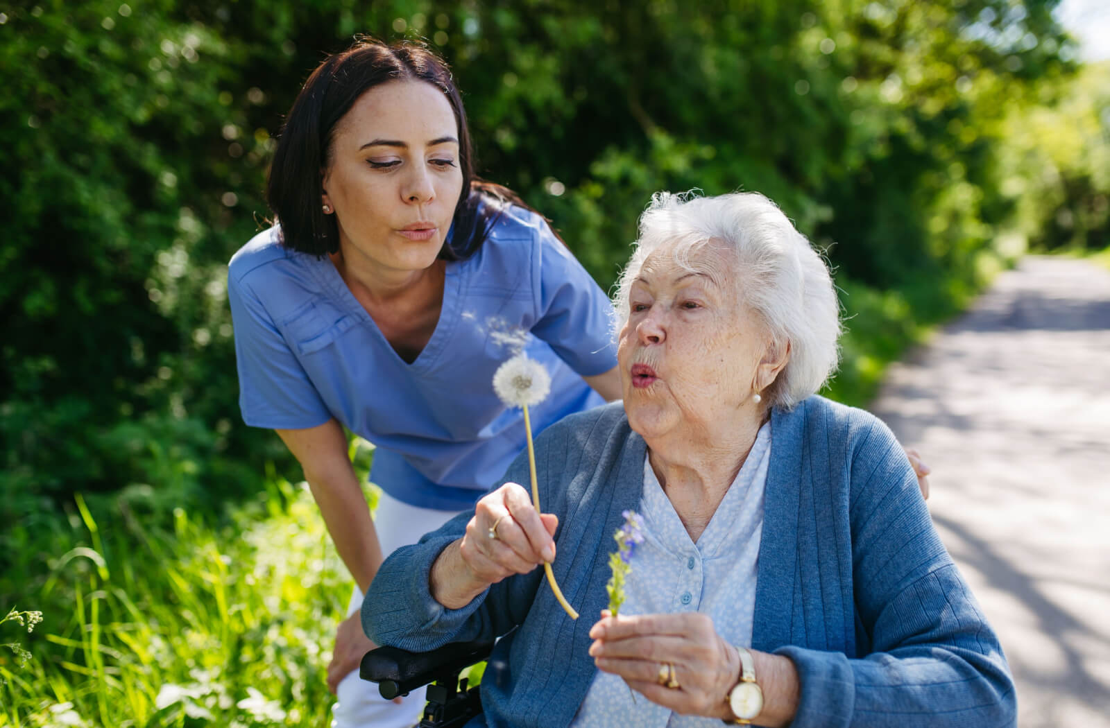 A caregiver pushing a resident in a wheelchair through a garden in senior living, blowing on a dandelion together.